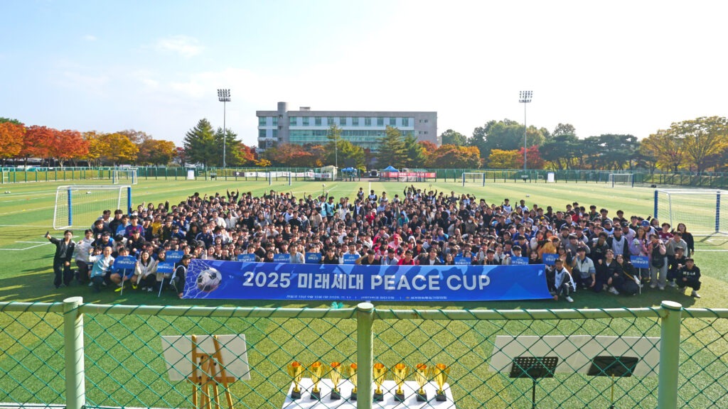 선문대학교 잔디구장에서 열린 ‘2025 신한국 미래세대 PEACE CUP’에 참가한 선수단과 참석자들이 대형 현수막을 중심으로 모여 손을 흔들며 단체 기념촬영을 하고 있다.